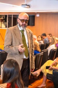 Um homem com barba, óculos escuros e terno marrom está em pé, no meio de um auditório, gesticulando enquanto conversa com a plateia.