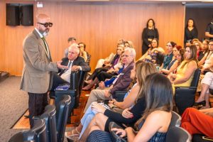 Um homem com óculos escuros e terno marrom está em pé, à frente de um grupo de pessoas sentadas em um auditório, e gesticula enquanto fala.