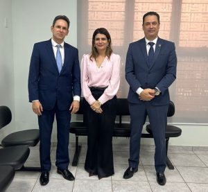 A imagem mostra um grupo de três pessoas, uma mulher e dois homens, em pé e posando para a câmera em um ambiente interno. Eles estão alinhados, com a mulher no centro. Os dois homens estão vestidos de terno e gravata, e a mulher usa uma blusa rosa clara e calça preta. O fundo tem persianas em uma janela e algumas cadeiras de couro pretas.