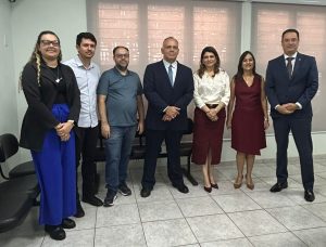 A imagem mostra um grupo de sete pessoas, cinco homens e duas mulheres, em pé e alinhadas para a foto em um ambiente de escritório. Todos estão em trajes de negócios ou social-casual e sorrindo para a câmera.