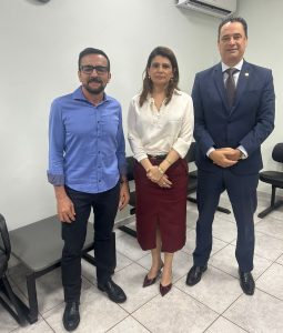 A imagem mostra um grupo de três pessoas, uma mulher e dois homens, em pé e posando para a câmera em um escritório. Da esquerda para a direita: um homem de óculos e camisa azul, uma mulher de blusa creme e saia vinho, e um homem de terno azul escuro. Eles estão em frente a uma parede branca, com cadeiras pretas e mesas de canto visíveis ao fundo.