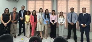 A imagem mostra um grupo de dez pessoas, cinco homens e cinco mulheres, em pé e posando para a câmera em um ambiente de escritório. Todos estão em trajes sociais ou profissionais. O grupo está alinhado, sorrindo e olhando para a câmera. Ao fundo, há uma grande janela com persianas, e no primeiro plano, mesas e cadeiras de escritório.