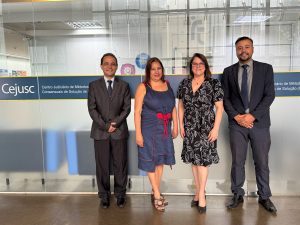 Foto colorida de quatro pessoas em pé em frente a portas de vidro. São dois homens usando terno e duas mulheres usando vestido. Ao fundo, colada na porta de vidro, está uma faixa azul escura com o texto "Cejusc - Centro Judiciário de Métodos Consensuais de Solução de Disputas".