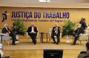 Três homens sentados no palco de um auditório que tem a logomarca da Justiça do Trabalho ao fundo