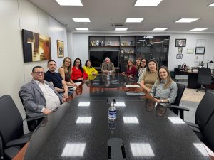 Várias pessoas sentadas ao redor de uma mesa oval. Atrás delas, uma estante preta vazada