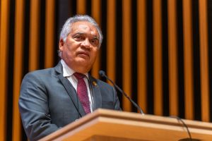 Um homem de cabelos grisalhos e terno está falando em um púlpito, com um microfone à sua frente. Ao fundo, uma parede com painéis de madeira.