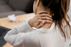 Mulher de camisa branca e cabelos presos está de costas apoiando a mão esquerda no pescoço