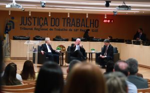 Três homens usando terno preto sentados em poltronas no palco de um auditório. Eles falam ao microfone.