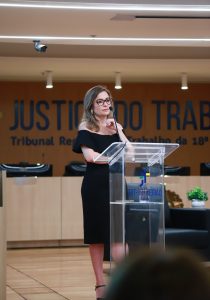 Mulher fala ao púlpito. Ela usa vestido preto e óculos