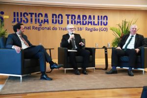 A imagem mostra três homens em trajes formais sentados em poltronas, participando de um painel ou conversa. O homem ao centro segura um microfone enquanto fala, e os outros dois sorriem e o escutam. Ao fundo, há um painel com a inscrição “Justiça do Trabalho – Tribunal Regional do Trabalho 18ª Região (GO)”.