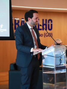 A imagem mostra um homem de terno escuro e gravata vermelha com padrões, falando em um púlpito de acrílico com microfone. O púlpito exibe o logotipo e o nome do TRT-18ª Região – Goiás. Ao fundo, aparece parte do letreiro “Justiça do Trabalho”.