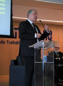 A imagem mostra um homem de terno escuro e gravata, falando em um púlpito de acrílico com microfone. Ele gesticula com a mão enquanto se dirige ao público. O púlpito traz o logotipo do TRT-18ª Região – Goiás, e ao fundo aparece parte do letreiro “Justiça do Trabalho”.