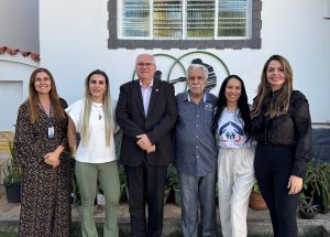 A imagem mostra um grupo de **seis pessoas** (quatro mulheres e dois homens) em pé e alinhados para uma foto, em frente a uma parede branca de um edifício. O grupo tem variados trajes, que vão do terno social a roupas casuais elegantes. Ao fundo, acima da cabeça do grupo, há uma **janela** retangular e um **logotipo decorativo** na parede.