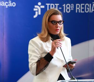 Imagem colorida de uma mulher de pele clara e cabelos loiros. Ela tem cabelo curto e está usando um óculos de grau com armação escura e um blazer branco. Com a mão esquerda ela segura um celular, enquanto a mão direita está apoiada em um microfone de mesa. Ao fundo, um painel na cor azul escuro com o símbolo da Justiça do Trabalho e o texto "TRT-18ª Região".