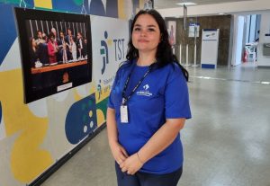 Uma mulher jovem de cabelos escuros e com uma camiseta azul com o logo da Justiça do Trabalho está de pé e posa para a câmera. Ao fundo, está uma parede com um quadro de foto e o logo do TRT-18ª Região.