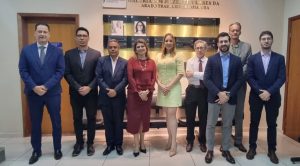 Um grupo de nove pessoas, sendo sete homens e duas mulheres, está em pé e posando para a foto em um corredor. Ao fundo, uma parede decorada com uma placa que diz "GALERIA DOS JUÍZES TITULARES DA VARA DO TRABALHO DE GOIATUBA" e fotos emolduradas.