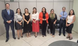 Um grupo de oito pessoas, incluindo um homem de terno e sete mulheres em trajes de negócios, posando lado a lado em um ambiente interno.