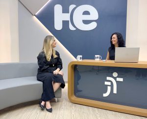 Uma mulher sentada em uma poltrona e um mulher numa bancada. Elas olham para a câmara. Ao fundo, a logomarca do programa hora extra. Embaixo da bancada, a logomarca da Justiça do Trabalho
