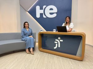 Uma mulher sentada em uma poltrona e um mulher numa bancada. Elas olham para a câmara. Ao fundo, a logomarca do programa hora extra. Embaixo da bancada, a logomarca da Justiça do Trabalho