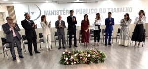 Autoridades durante solenidade no MPT-GO. São dez pessoas de pé, sendo cinco mulheres e cinco homens. Eles estão de pé, lado a lado. Ao fundo, uma parede branca com o texto "Ministério Público do Trabalho" em letras cinzas. No rodapé da imagem, um arranjo de flores.