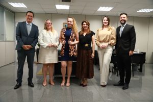 Três homens e três mulheres estão em pé, posando para uma foto em um ambiente interno. Todos estão vestidos em trajes sociais.