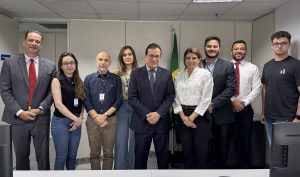 
Um grupo de nove pessoas, homens e mulheres, vestindo trajes de negócios e casuais, posando lado a lado em um escritório, com uma bandeira do Brasil ao fundo.