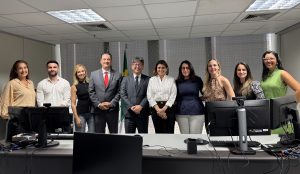 A imagem mostra cerca de dez pessoas, a maioria mulheres e dois homens, posando em fila atrás de uma mesa de escritório com monitores de computador. Todos estão vestidos com trajes sociais ou esporte fino e olham para a câmera. Ao centro, há um homem de terno cinza e uma mulher de camisa branca, ladeados pelos demais. Uma bandeira do Brasil é visível ao fundo, atrás do homem de terno.