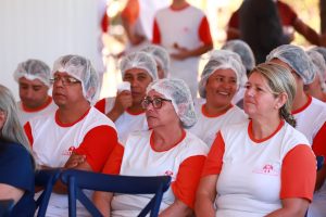 A imagem mostra um grupo de trabalhadores sentados e atentos, vestindo uniformes brancos com mangas vermelhas e toucas de proteção. Alguns sorriem, criando um clima de atenção e descontração durante o evento.