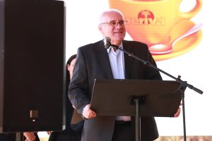 A imagem mostra um homem de terno e óculos falando ao microfone em um púlpito. Ele sorri enquanto se dirige ao público, com um telão ao fundo exibindo a imagem de uma xícara e o logotipo “Jerivá”.