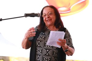 A imagem mostra uma mulher sorridente falando ao microfone enquanto segura um papel com anotações. Ela gesticula com uma das mãos, transmitindo simpatia e entusiasmo durante sua fala.