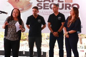 A imagem mostra quatro pessoas em pé diante de um painel do evento “Café Seguro”. Uma mulher à esquerda fala ao microfone segurando um papel, enquanto as outras três pessoas a observam atentamente, demonstrando interesse e atenção.