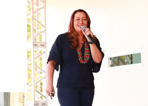 A imagem mostra uma mulher sorrindo enquanto fala ao microfone em um palco iluminado. Ela veste blusa escura com colar colorido e transmite simpatia e confiança durante sua apresentação.