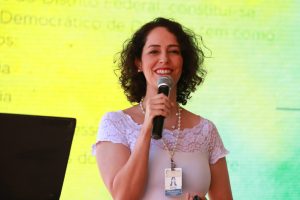 A imagem mostra uma mulher sorridente segurando um microfone enquanto fala diante de um fundo iluminado em tom amarelo. Ela usa blusa branca rendada e crachá, transmitindo simpatia e segurança durante a apresentação.