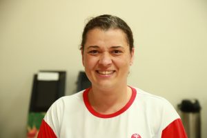 A imagem mostra uma mulher sorrindo para a câmera, vestindo uma camiseta branca com detalhes vermelhos. O fundo é neutro e levemente desfocado, destacando seu rosto expressivo e simpático.