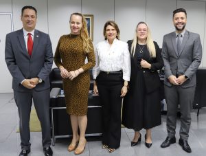 Um grupo de cinco pessoas, dois homens de terno e três mulheres com trajes de negócios e casuais elegantes, posando em pé em um escritório.