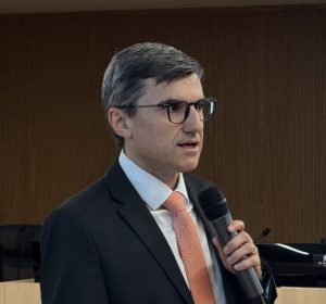 A imagem mostra um homem de meia-idade, vestindo terno e gravata cor de salmão, com óculos, discursando em um microfone. Ele está em um ambiente interno com painéis de madeira ao fundo, e parte de uma tela de projeção com texto é visível à direita. Ele olha levemente para o lado direito.