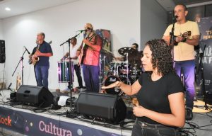 A imagem mostra uma performance musical em um palco. Em primeiro plano, uma mulher com cabelo cacheado e roupa preta faz a interpretação em libras. No plano de fundo, há cinco músicos no palco: um homem de camiseta amarela tocando violão e cantando, e outros músicos tocando bateria, saxofone e percussão. 