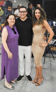 A imagem mostra três pessoas sorrindo para a câmera em um evento. No centro, um homem veste uma camiseta preta e calças brancas. À esquerda, uma mulher usa um vestido roxo longo e sapatilhas prateadas. À direita, uma mulher alta usa um vestido justo cor nude, sandálias de salto alto pretas e tem cabelo longo e cacheado. O fundo está ligeiramente desfocado, mostrando a área de um evento com banners e outras pessoas.