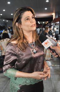 Uma mulher de pele clara e cabelos castanhos ondulados, vestindo uma blusa de cetim marrom-acinzentado com detalhes de renda verde e um colar com pingente de pedra grande, está sendo entrevistada. O ambiente de fundo está desfocado e é um evento interno com várias pessoas.