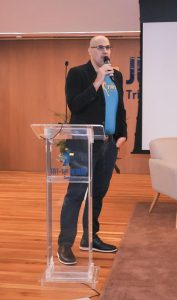 Homem branco, usando paletó e calça pretos, e camiseta azul clara.