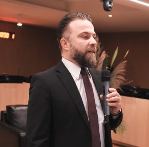 O orador, um homem de cabelo escuro e óculos, traj A imagem mostra um homem de meia-idade, vestindo terno e gravata, em pé atrás de um púlpito de acrílico transparente. Ele está discursando e olhando para frente. O púlpito exibe o logotipo do "TRT - 18ª Região Goiás". O ambiente parece ser um auditório ou sala de sessões, com piso de madeira, um painel marrom e bandeiras visíveis ao fundo.