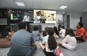 Foto de crianças durante visita à unidade de TI do TRT-GO. Elas estão sentadas no chão, de frente a duas telas grandes e assistem a explicação de um servidor sobre a unidade.