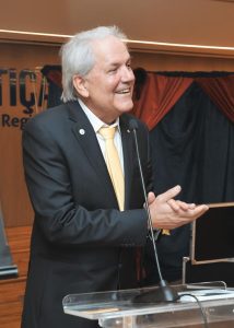 Foto de um homem de pele clara e cabelos grisalhos. Ele usa um terno preto com gravata amarela e está de pé, em frente a um púlpito e sorrindo.