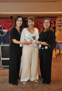 Foto de três mulheres sorrindo e posando para foto enquanto seguram um livro de colorir. Elas têm pele clara e usam roupas sociais.