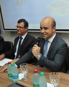 Foto de um homem de pele clara e cabelos escuros. Ele está usa um terno escuro com gravata azul. Ele está sentado e falando ao microfone.