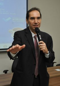 Foto de um homem de pele clara e cabelos escuros. Eles está usando um terno preto com gravata rosa, e está falando ao microfone.