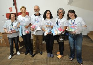 Um grupo de seis pessoas, incluindo um homem e cinco mulheres, está em pé, posando para a foto. Todos usam camisetas brancas com a inscrição "POR VOCÊ, POR NÓS, POR UM JUD" e seguram sacolas transparentes com itens de higiene e cuidados pessoais.