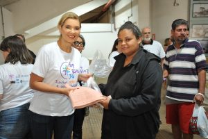 Uma mulher de camiseta branca com a estampa "POR VOCÊ, POR NÓS, POR UM JUD" entrega um kit de higiene embalado em plástico para outra mulher. Outras pessoas, incluindo um homem de barba, estão ao redor.
