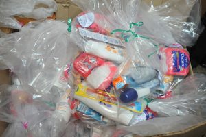 Uma caixa de papelão cheia de sacolas plásticas transparentes, recheadas com itens de higiene e cuidados pessoais como sabonetes, pastas de dente e loções.