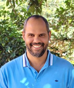 Homem de pele clara e cabelos escuros. Ele usa uma camisa azul claro. Atrás dele estão várias árvores.